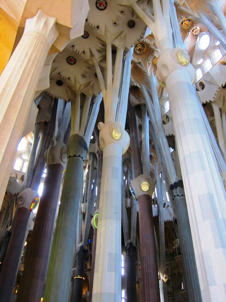 Very tall pale, green and reddish stone columns split into angled branches beneath a bright ceiling with flower-like openings.