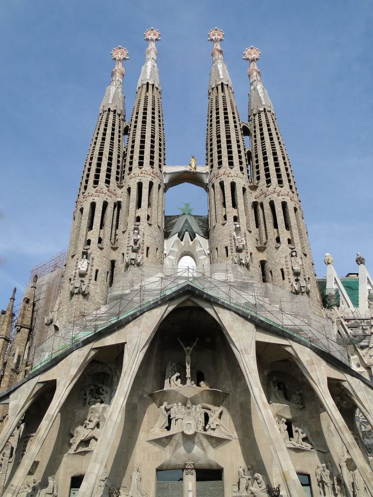 Four slender perforated towers rise above a stark stone façade with inclined porch supports, angular sculptures and scaffolding.