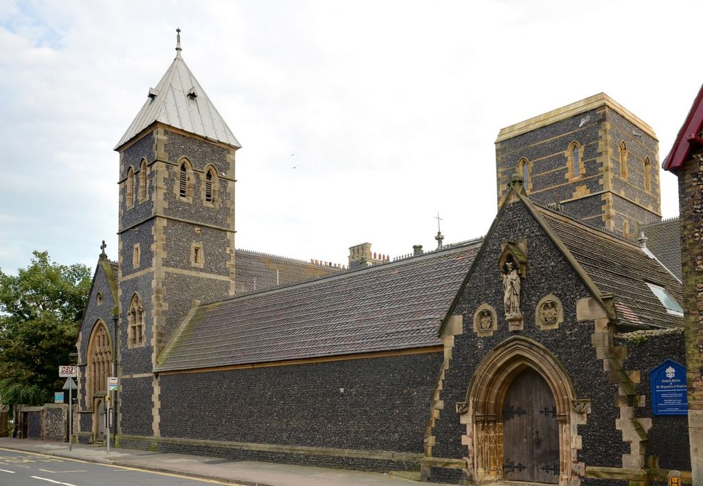 A long dark-flint Gothic Revival church complex lines a street, with pale stone around pointed doors and windows, steep tiled roofs, one tall spired tower and another square tower.