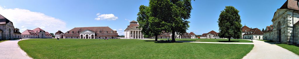 A very wide panorama shows pale stone saltworks buildings curving around a green court, with a larger pedimented director's house at the centre and trees breaking the view.