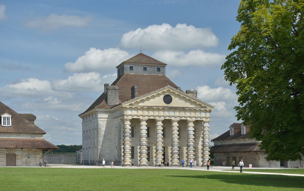 A monumental pale stone director's house stands in a grassy court, with a triangular pediment, steep roof and six unusual columns made from alternating round and square blocks.