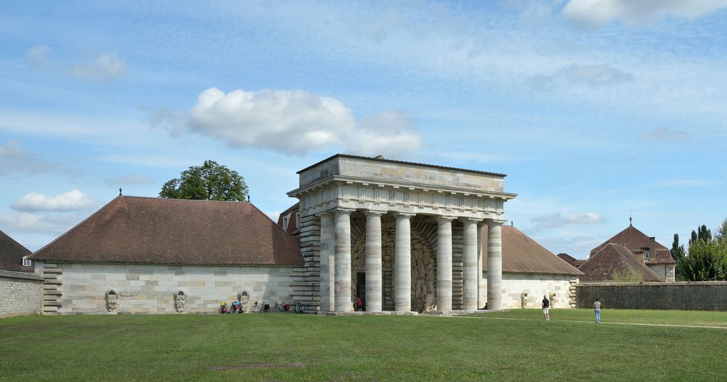 A long pale stone entrance building has six tall columns around a central passage lined with rough rock-like blocks, flanked by low hipped-roof wings across a lawn.