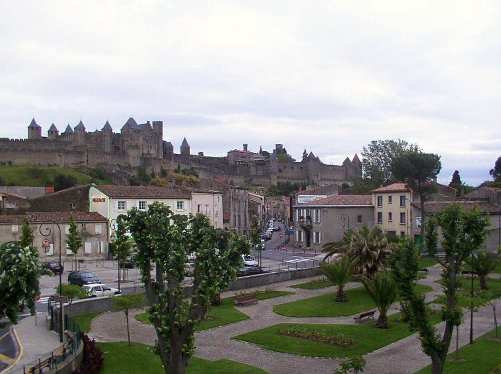 A long fortified hilltop city with double stone walls, many towers and steep conical roofs rises above modern streets, houses, gardens and clipped trees.