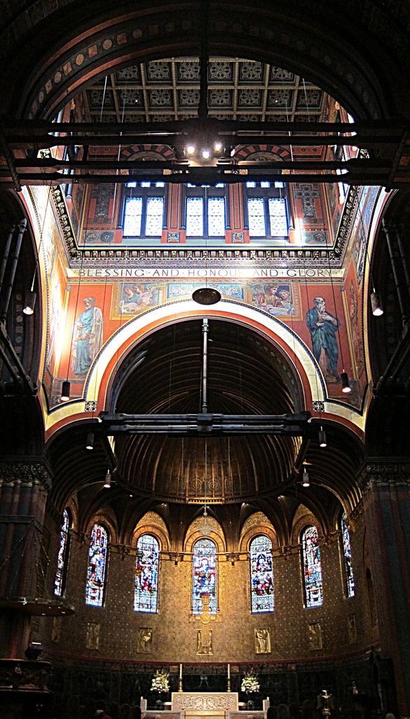 A tall church interior rises through a broad painted arch and dark timber beams to bright upper windows, with murals, patterned bands and stained glass around a curved sanctuary.