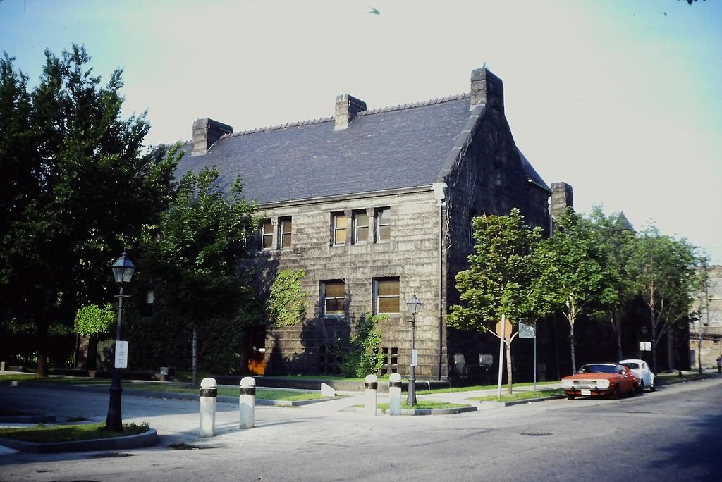 A dark rock-faced masonry house with a steep roof, tall chimneys and few street windows stands behind trees at a quiet Chicago corner.