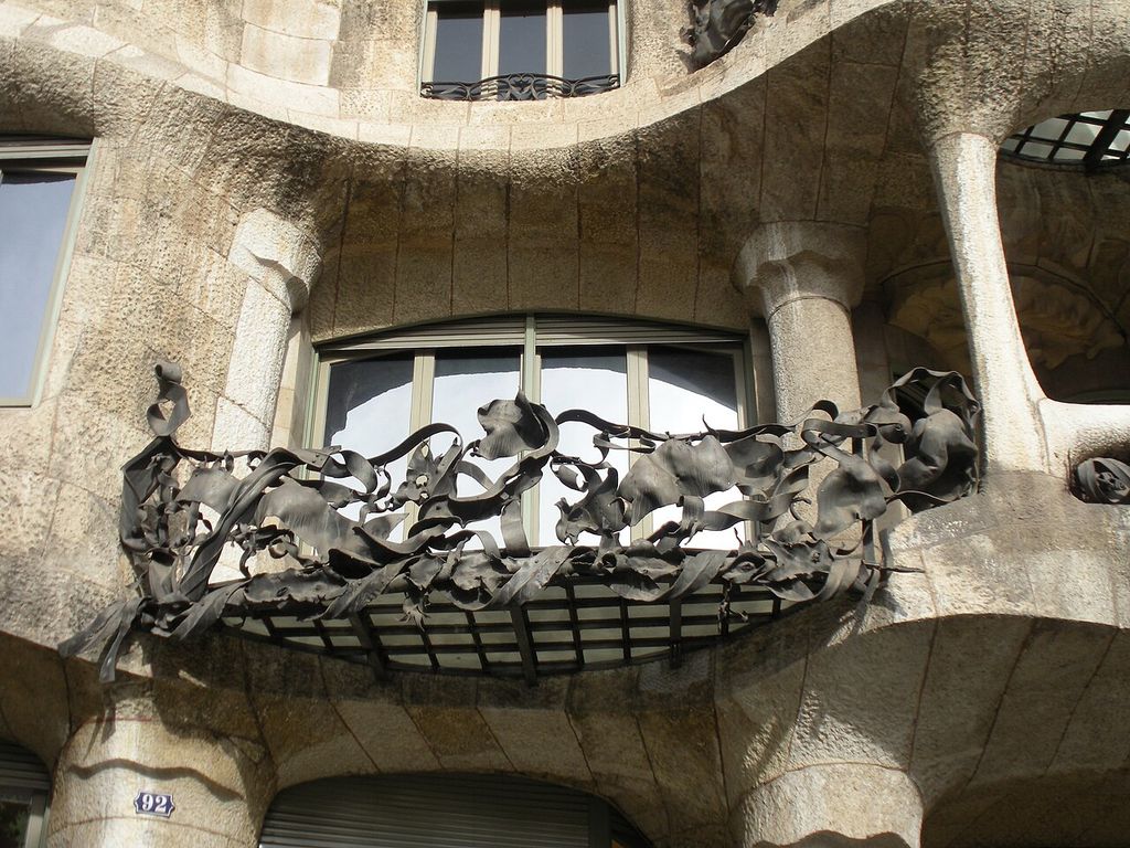 Irregular dark wrought iron shaped like curling leaves spans a balcony set between rough curving stone piers and rounded window openings.
