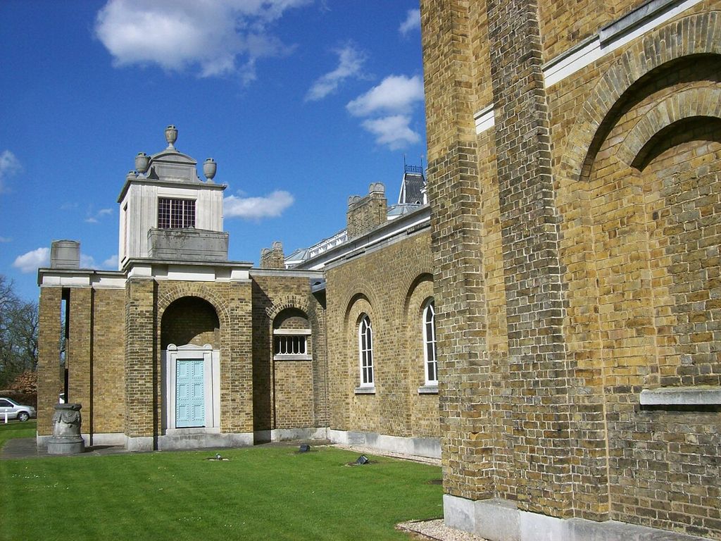 A low brick gallery wall with blind arches leads towards a taller pale-topped mausoleum block beneath a bright blue sky; a small parked car appears at the far left.