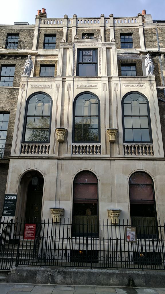 A narrow pale-stone townhouse front with three tall arched windows, abstract classical details, statues and a small upper loggia projects between darker brick buildings.