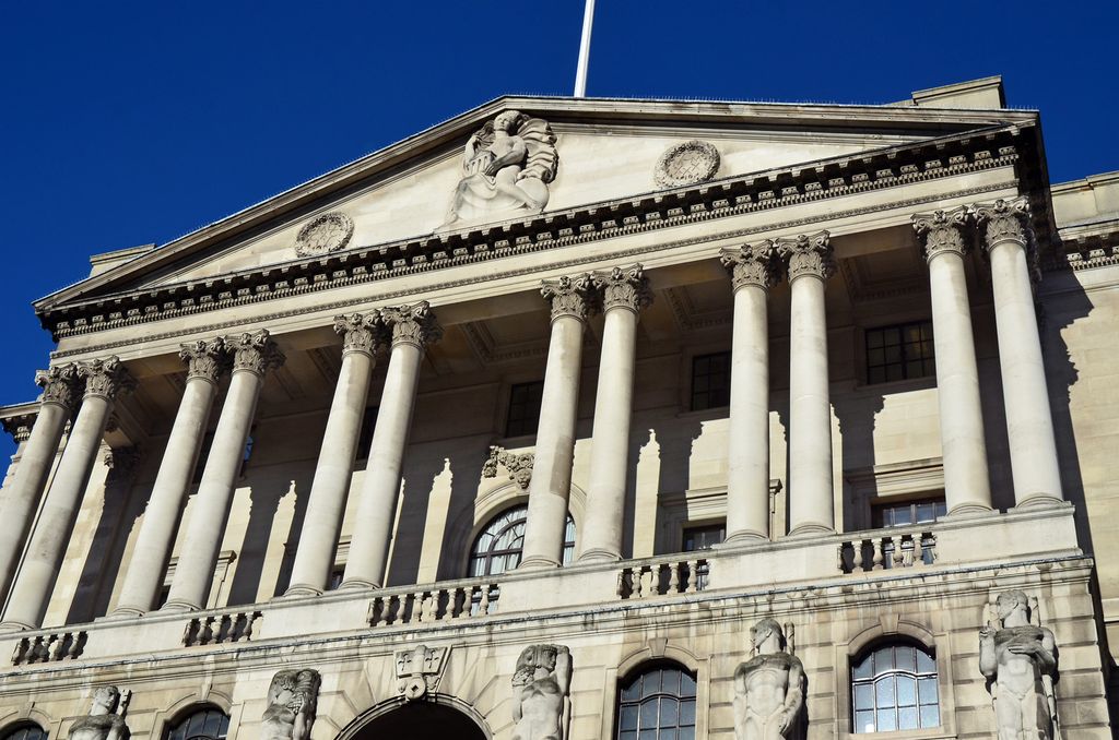 A low-angle close view shows a monumental pale-stone bank façade with tall Corinthian columns, a broad sculpted pediment and arched windows against a clear blue sky.