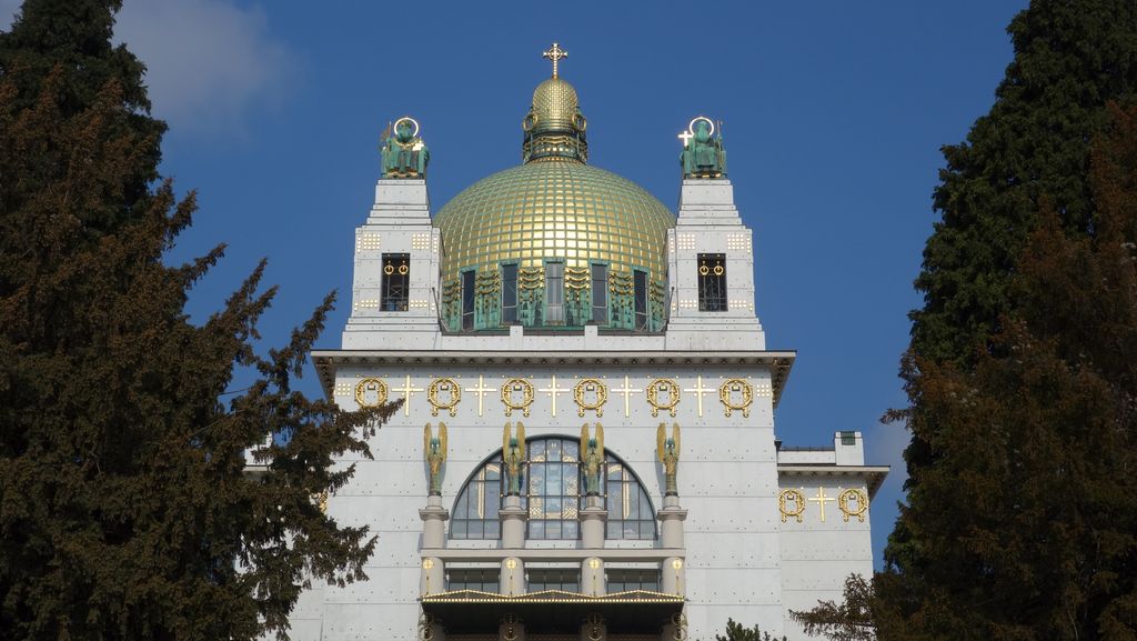 A frontal white church façade rises between dark trees to a large gold dome, with twin towers, green statues and bands of gold ornament around an arched central window.