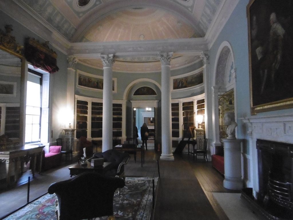 A pale blue library interior has tall paired columns, curved bookcases and painted panels beneath a coved ceiling, with a doorway continuing the axis through another room.