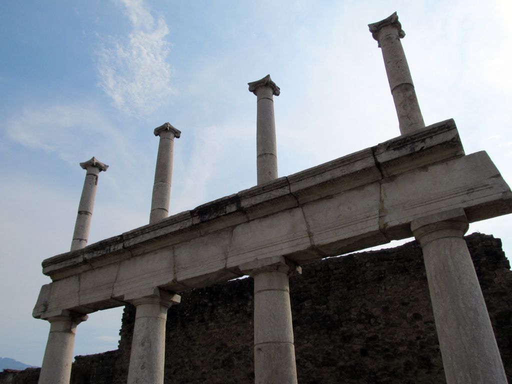 Five tall pale columns and capitals rise above a heavy lintel line before dark ruin walls, seen from a low angle against clouded sky.