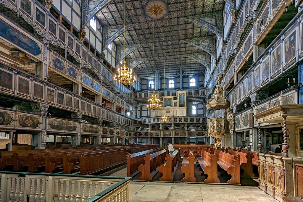 A wide timber-framed worship room is surrounded by richly painted gallery tiers beneath a decorated ceiling, with pews, organ, pulpit and chandeliers visible.