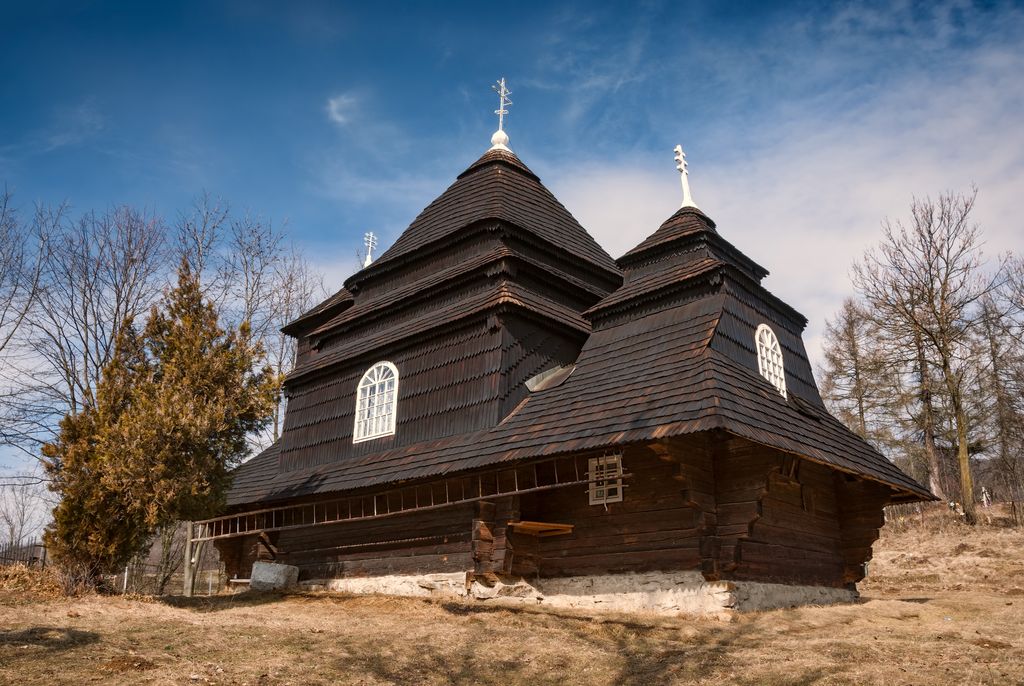 A low log-built church rises through three shingled roof masses and small crosses, with broad eaves, white-framed windows and leafless trees around it.