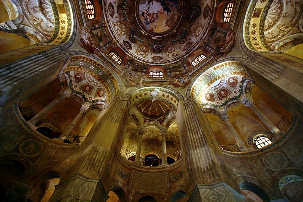 An upward wide-angle view passes tall piers and curved gallery arcades toward a richly decorated central dome, with lower areas in deep shadow.