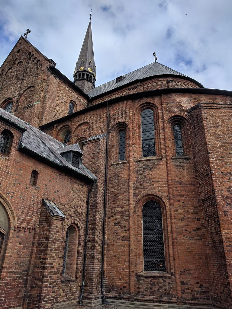 A close exterior view shows curving brick walls, round-headed windows, vertical strips, overlapping metal roofs and a slender spire against cloud.