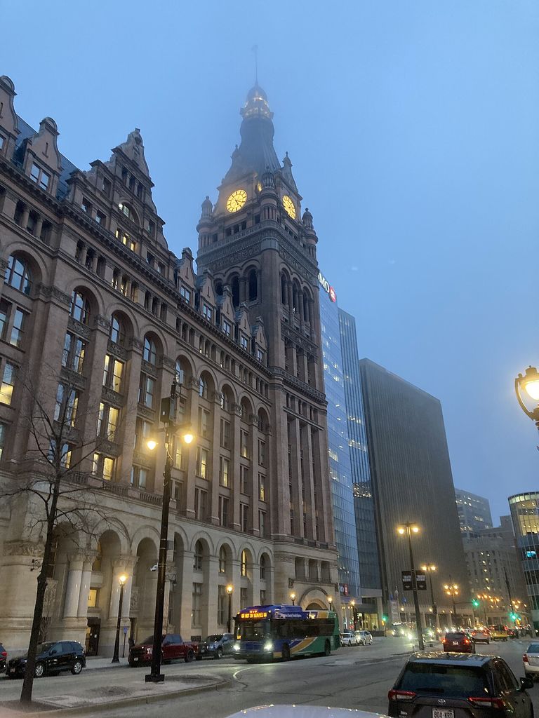 A tall historic masonry city hall with arched windows and glowing clock faces rises through fog beside glass towers, buses and cars.
