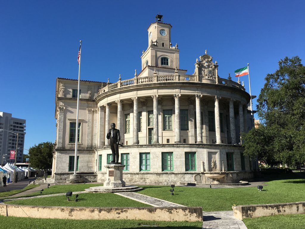A pale curved city hall with a columned upper loggia and square clock tower stands behind lawn, palms, flags and a bronze statue.