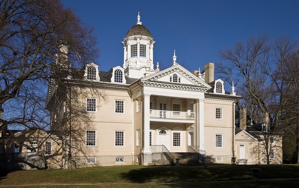 A pale country house has a tall columned portico, triangular pediment, cupola and lower flanking ranges partly screened by bare trees.