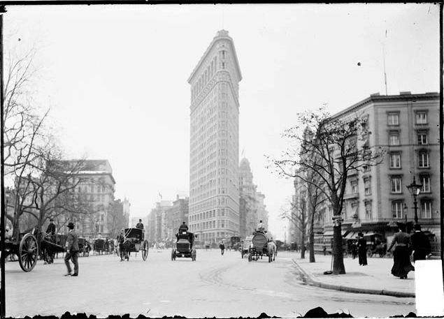 A historic city view shows the narrow wedge-shaped Flatiron Building rising behind a broad street filled with horse-drawn vehicles and pedestrians.