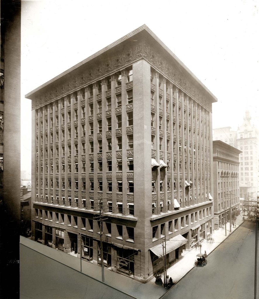 An elevated historical view shows a tall rectangular office block with a two-storey base, repeated narrow bays, deep projecting cornice, awnings and a horse-drawn vehicle.