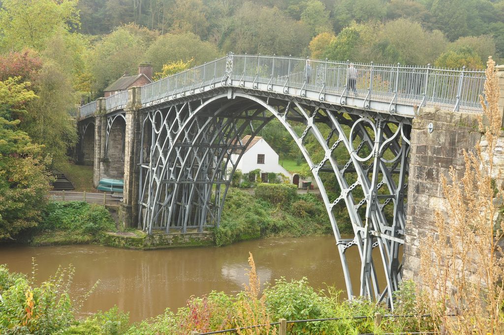A pale-grey iron arch with open lattice ribs spans a brown river between high stone abutments amid autumn trees.