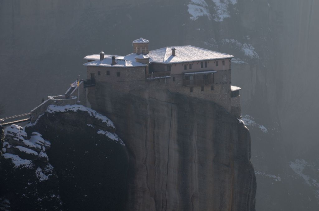 A compact roofed monastery rests on the sheer top of a tall rock pinnacle, with snow on ledges and a hazy cliff rising behind.