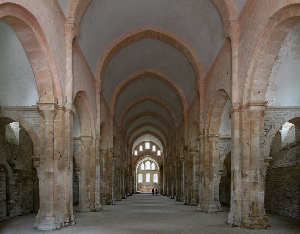 Austere pale-stone arcades and pointed transverse arches repeat along a high vaulted nave toward bright eastern windows.