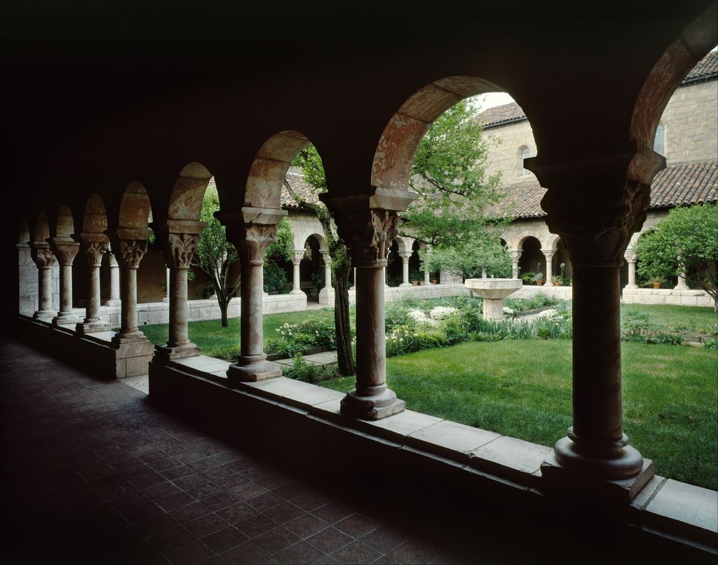 A dark arcaded gallery with carved capitals frames a sunlit planted court and fountain within a museum reconstruction.