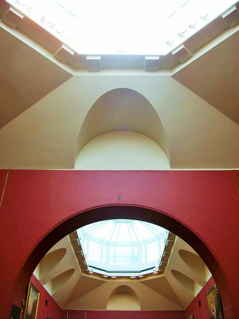 A bright rooflight illuminates shaped pale ceilings and a deep arch between red-walled picture galleries.