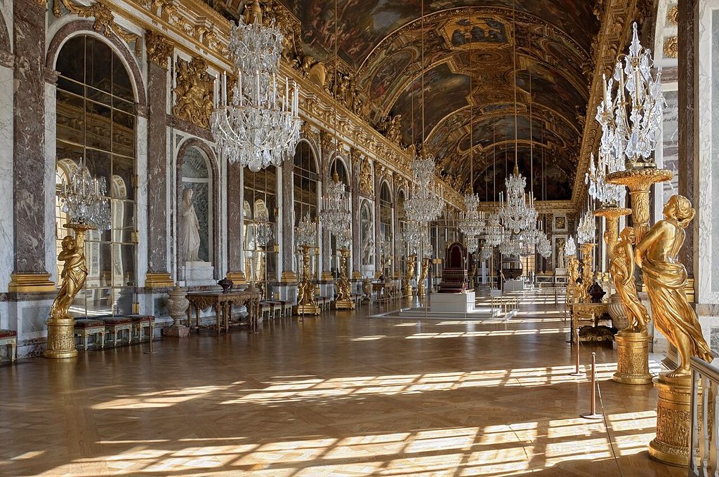A long gilded gallery aligns mirrored arches, marble pilasters, chandeliers and a painted barrel vault above sunlit parquet.