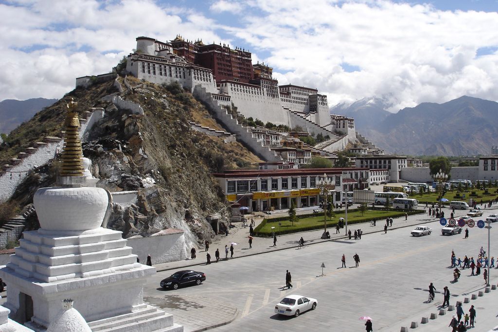A vast white and red palace climbs a rocky hill above Lhasa, with stepped walls, stairs, religious structures, roads, vehicles and mountains visible.