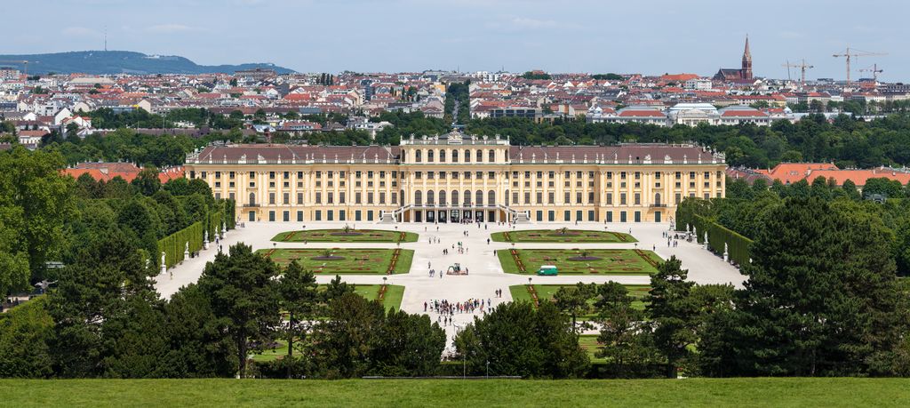 A long pale imperial palace faces symmetrical parterres and tree-lined paths, with Vienna extending across the horizon behind it.