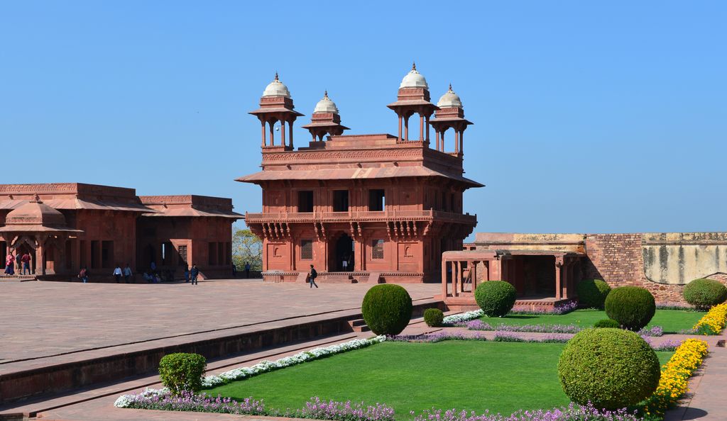 A square red-sandstone pavilion with deep eaves and four domed corner kiosks stands beside a court and clipped garden under blue sky.