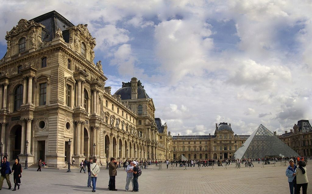 Ornate stone palace pavilions and arcades enclose a large court containing the modern glass pyramid beneath a clouded sky.