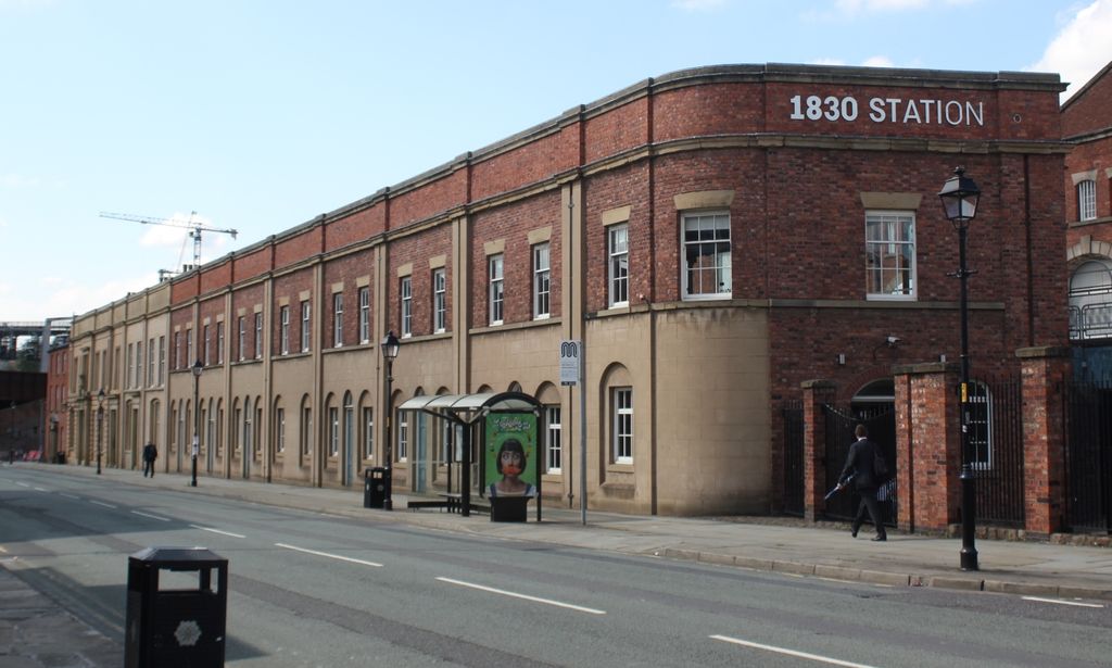 A long two-storey brick and pale-stone street range with repeated arched openings carries a modern sign reading 1830 STATION above a road and bus shelter.