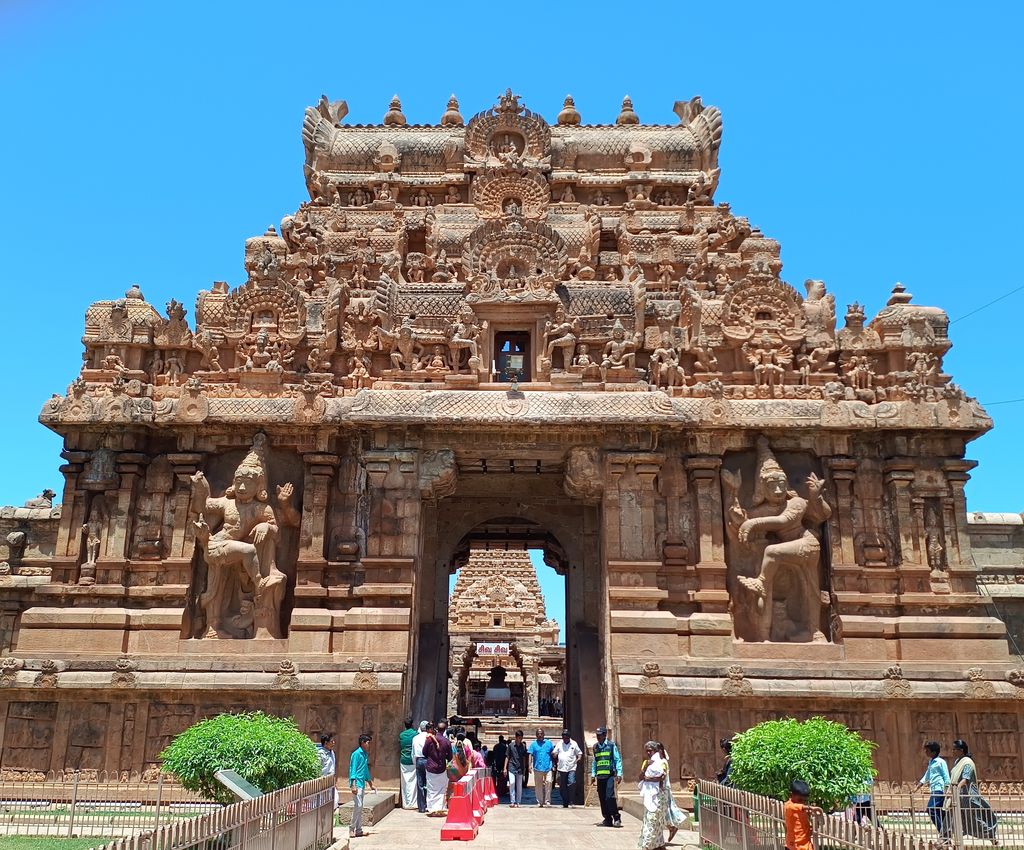 A richly sculpted stone gateway with large guardians frames a second tower and buildings deeper within the busy temple precinct.