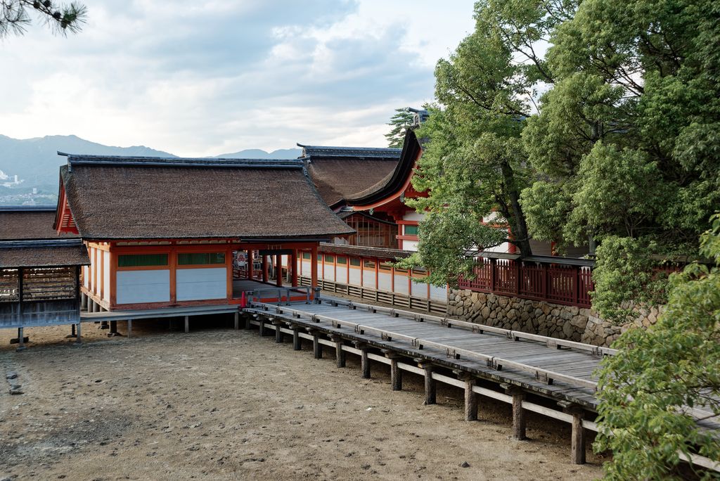 Red-and-white timber shrine buildings and a raised boardwalk cross an exposed tidal shore below trees and mountains.