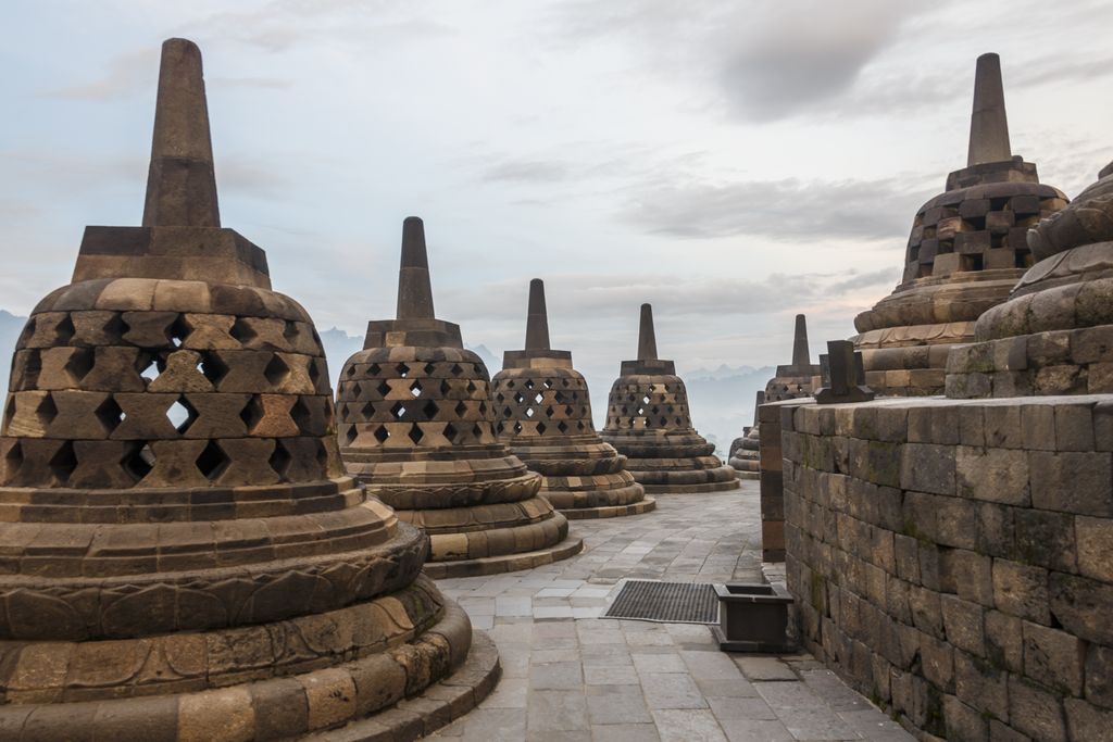 A stone path curves between rows of perforated bell-shaped stupas on an upper terrace, with hazy mountains beneath a cloudy sky.