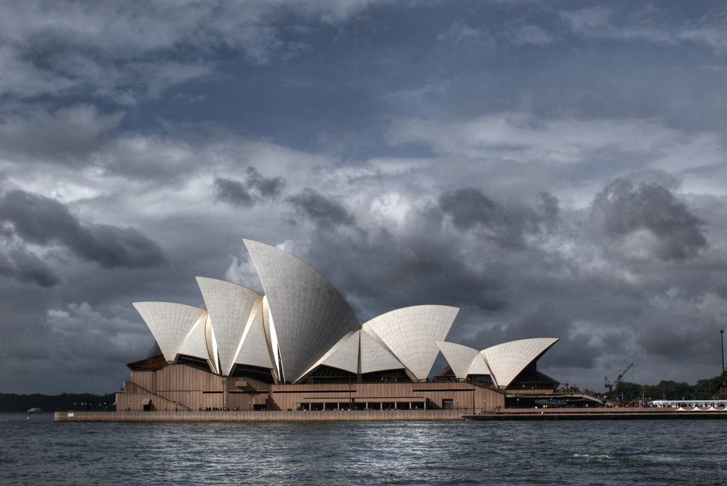 Multiple white shell roofs rise from a broad podium beside dark harbour water beneath heavy clouds.