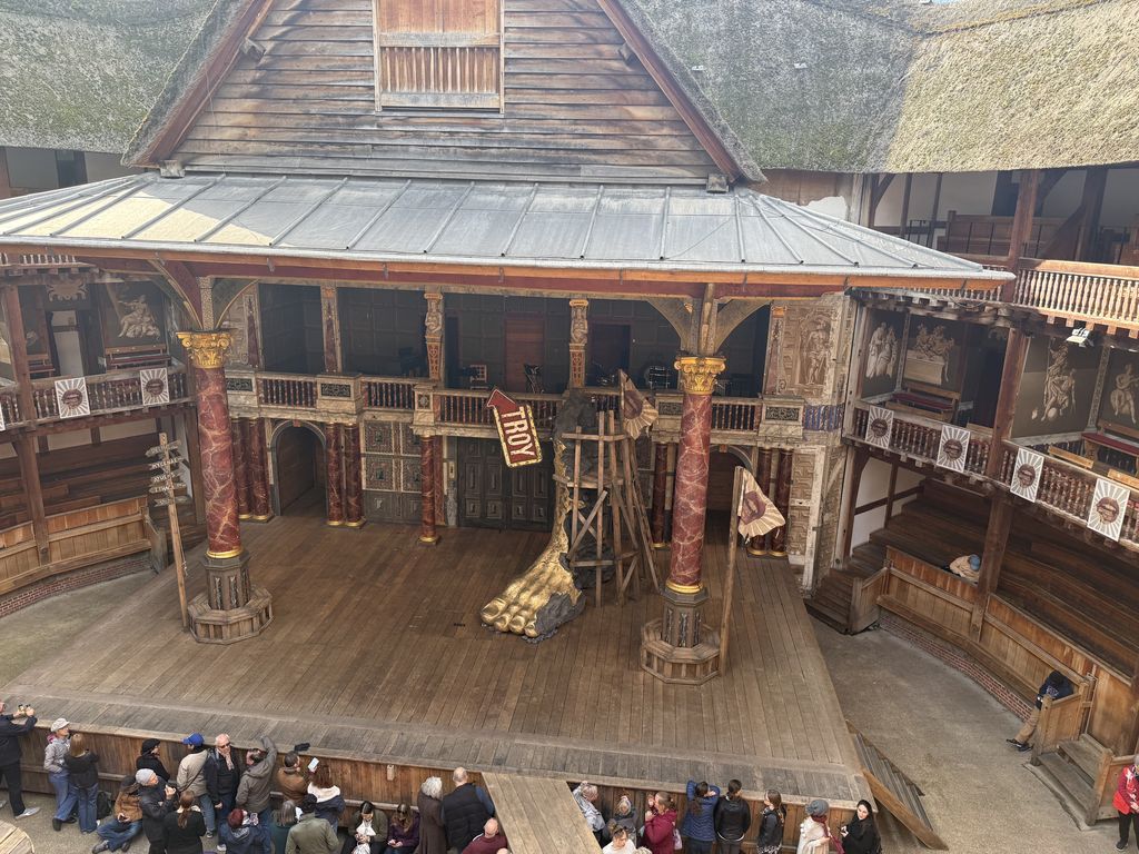 A decorated timber stage projects into an open yard enclosed by several tiers of covered wooden galleries and thatched roofs, with visitors and a temporary prop.
