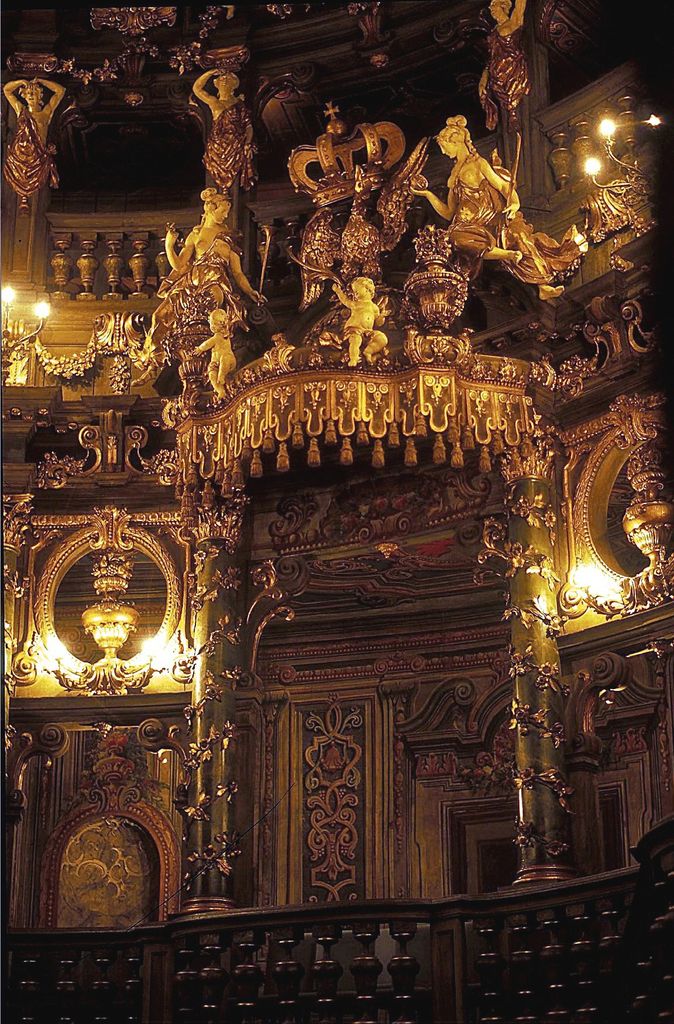 An elaborate gilded and dark-painted royal box carries a large crown, sculpted figures, lamps, columns and layered balcony rails.
