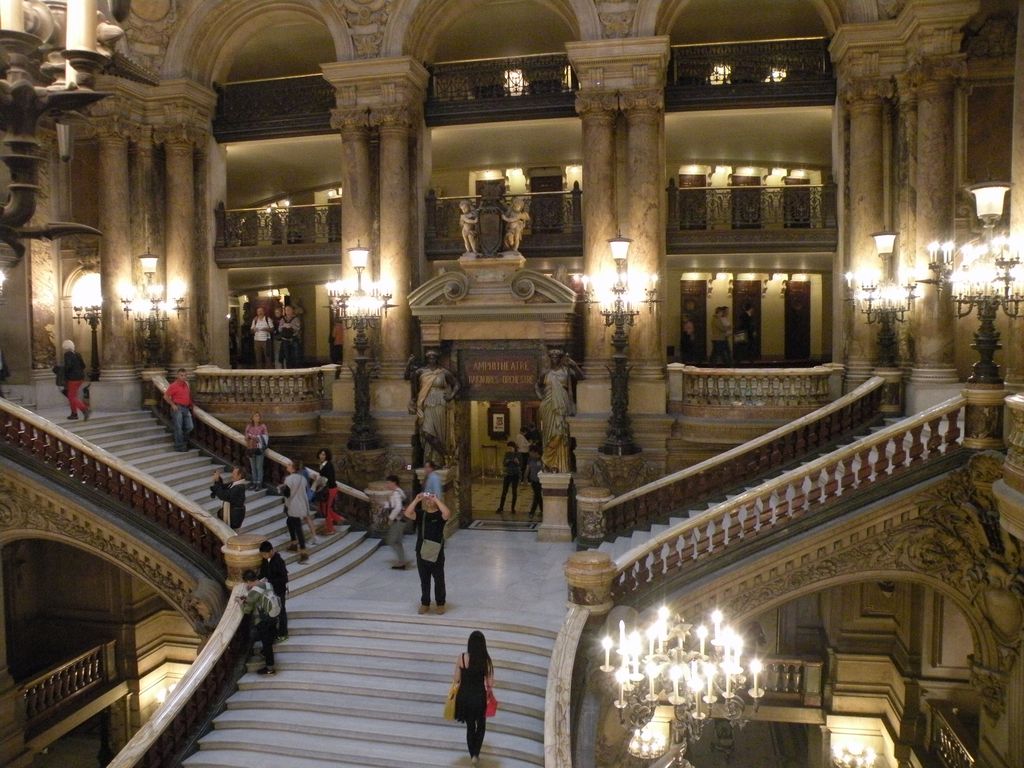 Branching marble stairs rise through a richly decorated hall of columns, arches, statues, lamps, balconies and moving visitors.