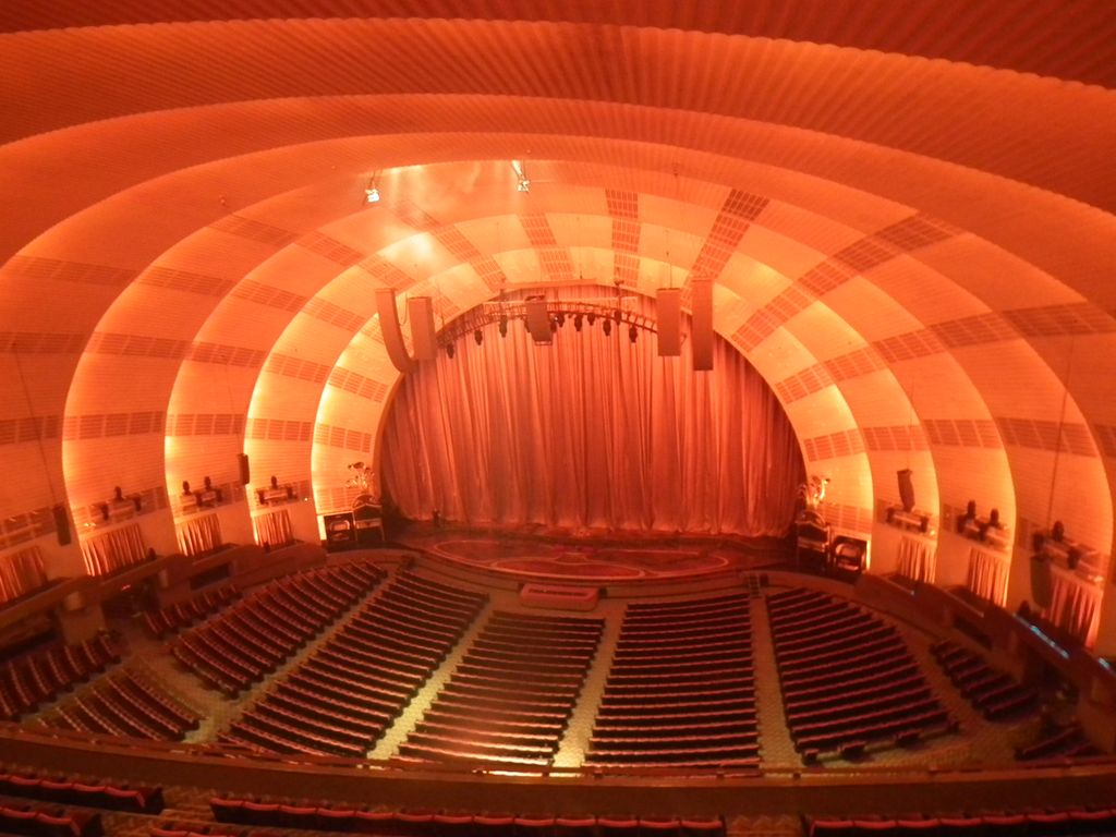 Rows of seats face a curtained stage beneath broad concentric ceiling arches glowing orange, with aisles and technical fixtures visible.