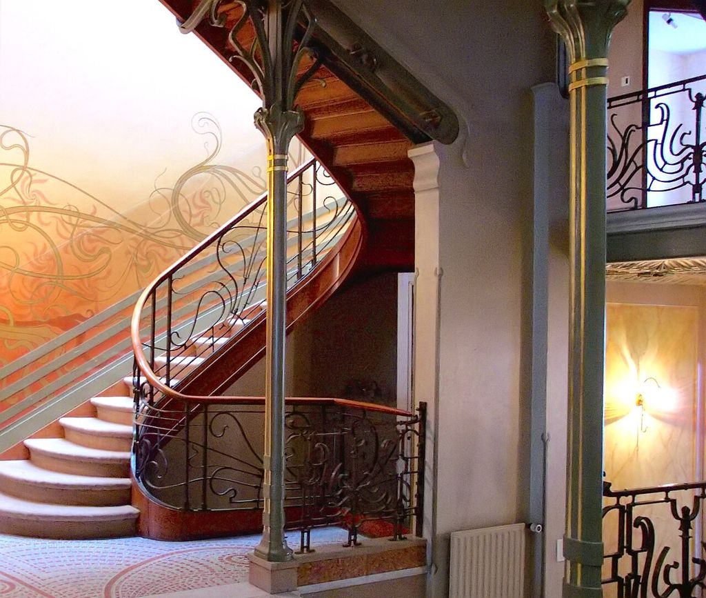 A curving stair and flowing iron rails rise among slender green metal columns, patterned floor, painted tendrils and glowing wall lights.