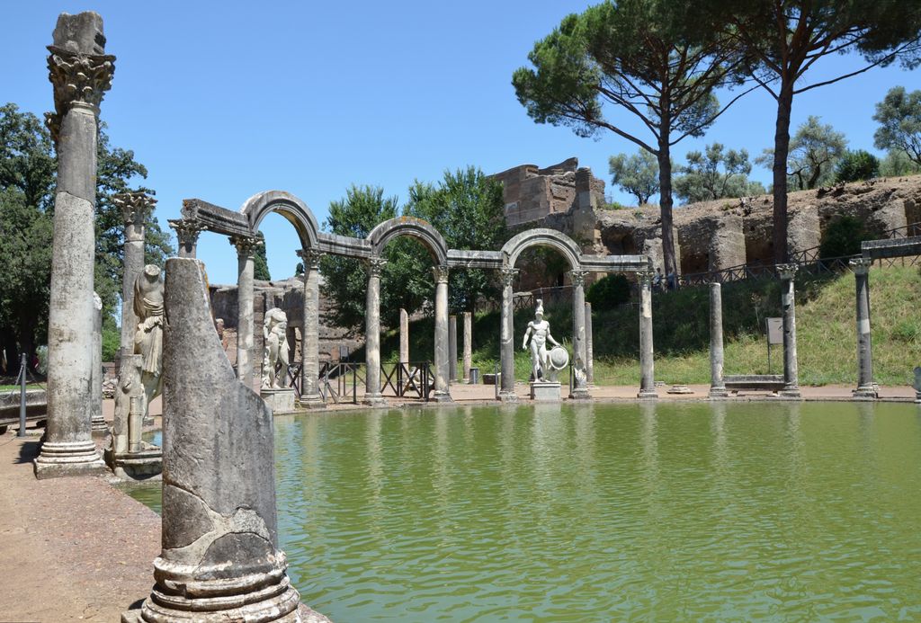 A long green pool faces a curved colonnade of columns and arches with statues, ruins, paths, railings and trees around it.