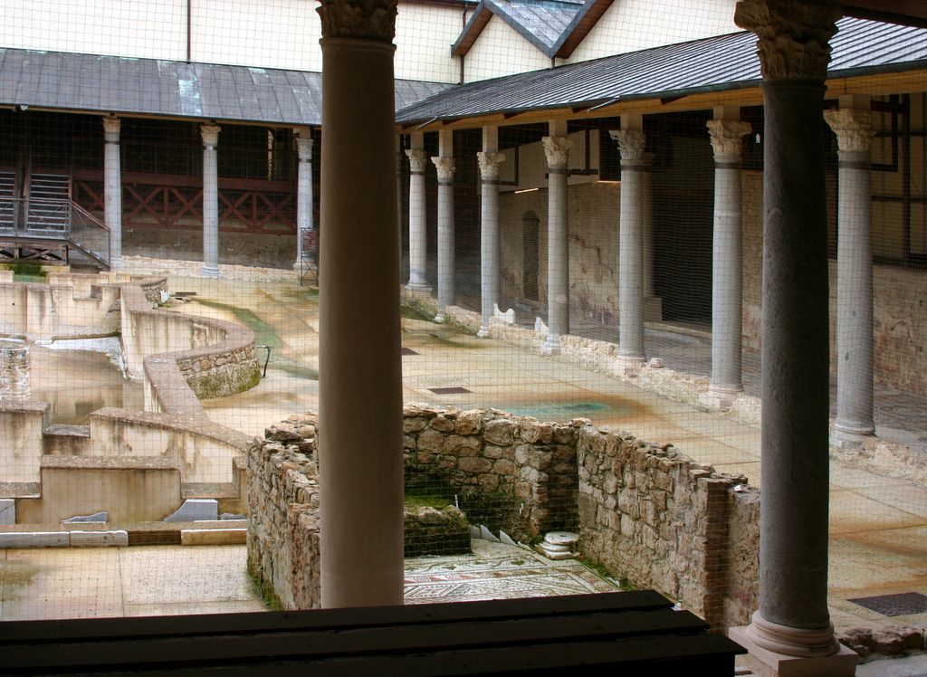 Ancient columns and mosaic floors surround a sunken peristyle fountain beneath modern protective roofs, netting and viewing structures.