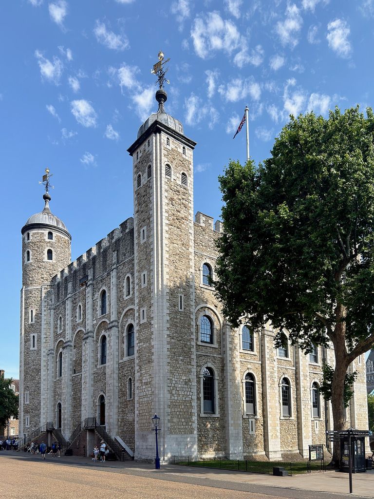 A tall pale-stone keep with corner turrets, small windows and steep later roofs rises behind a tree and visitor barriers.