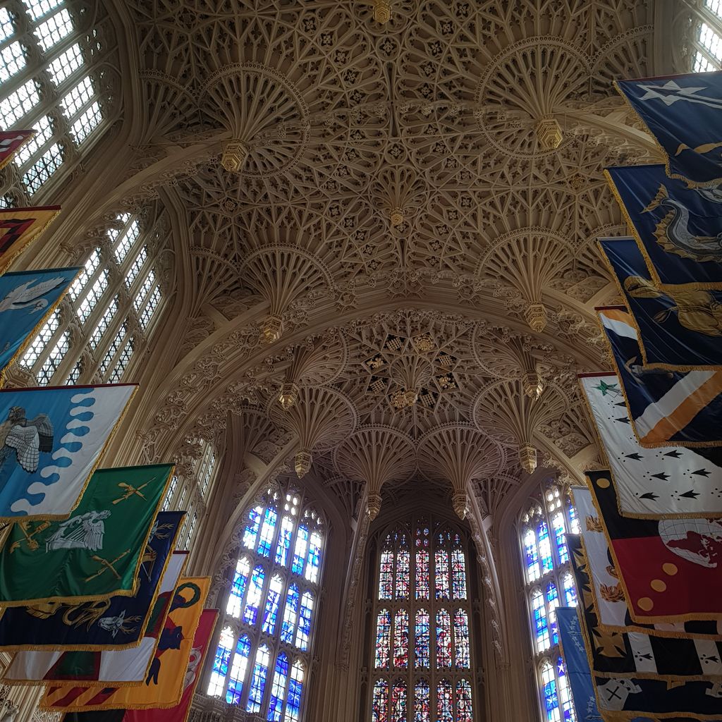 Dense pale stone fan vaults and hanging pendants cover a chapel ceiling above tall windows, with colourful ceremonial banners along both sides.