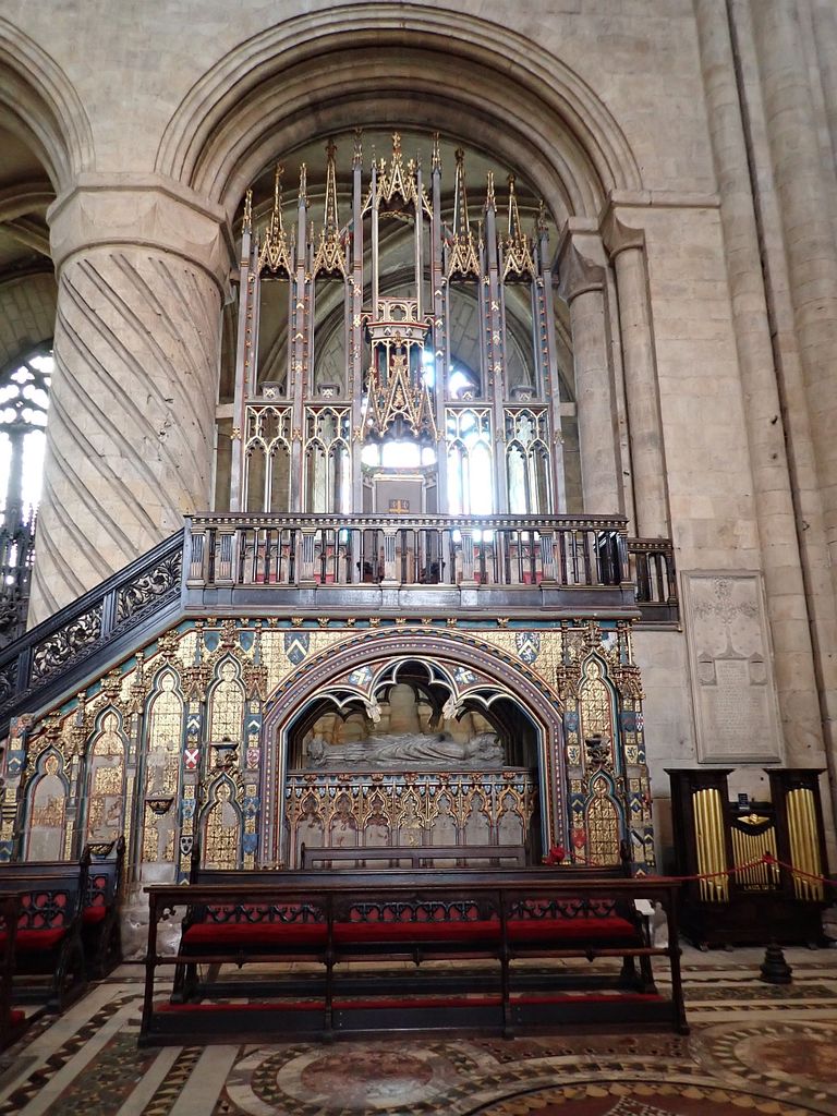 A raised seat and tall carved canopy stand behind a stone balustrade and stair above a canopied tomb inside Durham Cathedral.